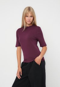 Une femme aux cheveux raides et blonds porte un haut violet à manches courtes et un pantalon noir à jambes larges, posant avec une main dans la poche sur fond blanc.