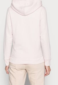 Sudadera con capucha de color rosa claro, usada por una persona vista desde atrás, combinada con pantalones beige sobre un fondo claro y liso.