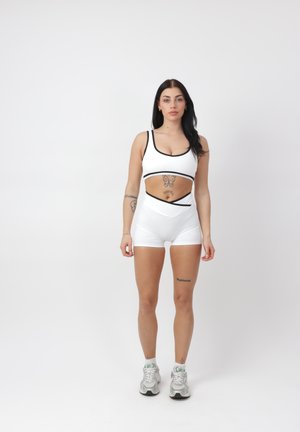 Frau steht in weißer Sportbekleidung und Turnschuhen, blickt nach vorn vor einem schlichten hellen Hintergrund, sichtbare Tattoos an Armen und Oberkörper.