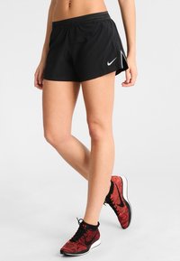 Pantalones cortos deportivos negros con una cinturilla ajustada, aberturas laterales y un pequeño logo blanco de Nike. Combinados con zapatillas de correr de patrón rojo y negro.