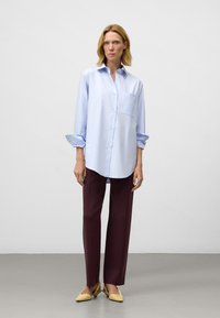 Lichtblauw overhemd met lange mouwen en een zak, gecombineerd met bordeauxrode rechttoe-rechtaan broek en gele ballerina's. Glad stoftextuur.