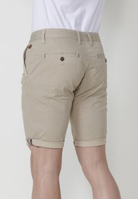 Beige woven shorts with a slim fit, featuring rolled hems, back pockets with buttons, and a textured finish.