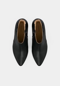 Une paire de bottines noires en cuir à bout pointu, avec des zips latéraux et une doublure intérieure beige, vues de dessus sur un fond blanc.