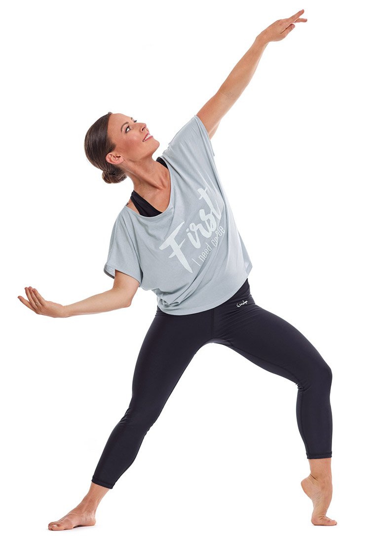 Camiseta de corte holgado en azul claro con texto blanco que dice "Primero necesito amor", combinada con leggings negros. La modelo está en una pose dinámica.