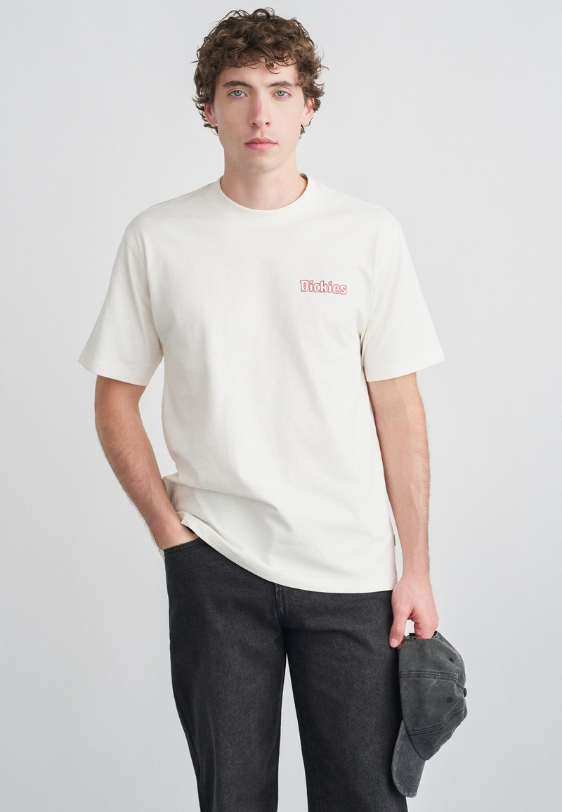 Kremhvid T-shirt med korte ærmer og lyserød "Dickies"-logo på venstre bryst, kombineret med mørke jeans og holder en grå kasket.