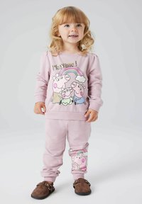 Gaiši rozā kokvilnas džemperis ar multfilmu cūkām un varavīksni, kombinēts ar atbilstošiem rozā biksēm ar rakstura dizainu uz kreisās kājas.