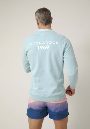 Hombre con sudadera azul clara con "ALTONADOCK 1969" en la espalda y pantalones cortos multicolor con patrón degradado azul y rosa, de espaldas.