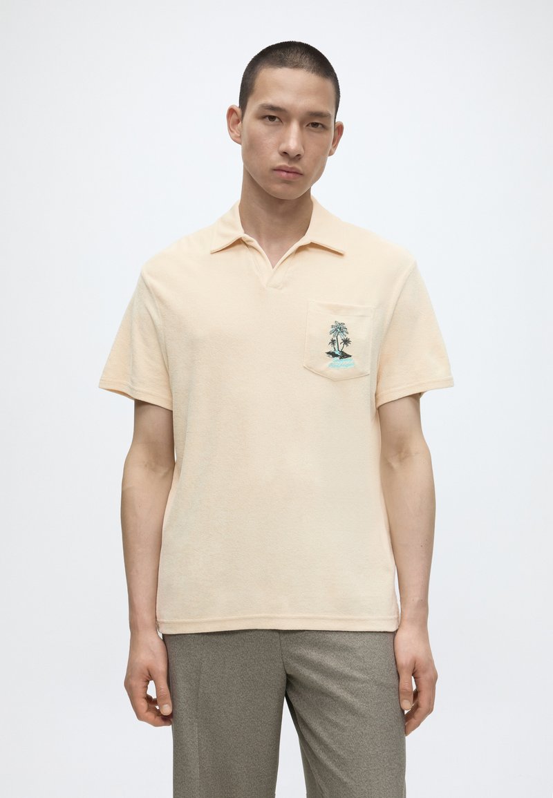 Jeune homme portant un polo beige à manches courtes avec un motif de palmier sur la poche poitrine et un pantalon gris, debout devant un fond blanc.