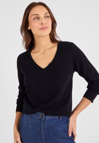 Zwarte v-hals trui met lange mouwen, gemaakt van zacht materiaal, met ribgebreide manchetten en zoom, gedragen met blauwe denim jeans.