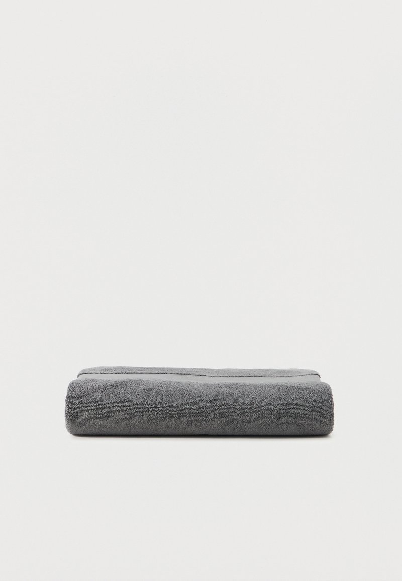 Διπλωμένη πετσέτα μπάνιου σκούρου γκρι χρώματος τοποθετημένη σε απλή λευκή επιφάνεια, με εμφανή την απαλή υφή από πετσέτα βελουτέ.