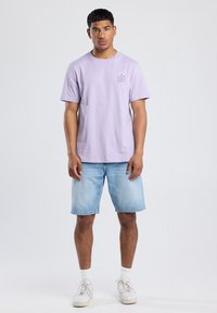 Vaalean violetti puuvillapaita, jossa on pieni musta kukkakuva rinnassa, yhdistettynä vaaleansinisiin farkkuhousuihin ja valkoisiin lenkkareihin.