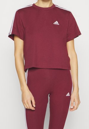 Osoba ubrana w bordowy krótki t-shirt Adidas oraz legginsy z białymi paskami na rękawach i bokach spodni, z logo Adidas na klatce piersiowej i udzie.