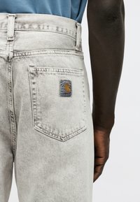 Világosszürke farmernadrág Carhartt logóval a hátsó zsebén, amelyet egy sötét bőrű, kék inget viselő személy hord.