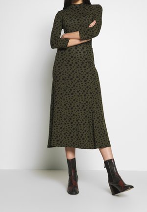 Kobieta ubrana w oliwkowozieloną, długą sukienkę o długości do połowy łydki z czarnymi cętkami w kształcie lamparta oraz czarne botki z czerwonymi akcentami, stojąca z założonymi ramionami.