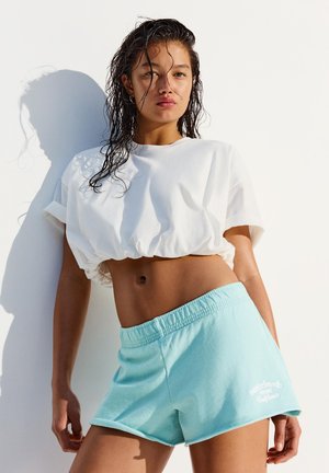 Giovane donna con capelli bagnati, indossa una t-shirt bianca corta e pantaloncini azzurri chiari, in piedi contro un muro bianco alla luce del sole.