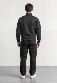 Musta bomberi takki ja tekstuurilla valmistetut housut, joissa on ribattu hihansuu ja vyötärö, yhdistettynä valkoisiin tennareihin, nähtynä takaa.