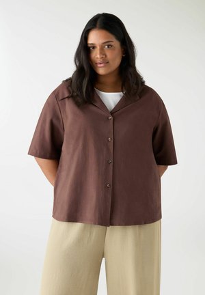 Donna con capelli castani di media lunghezza che indossa una camicia marrone a maniche corte con bottoni e pantaloni beige chiaro, davanti a uno sfondo semplice.