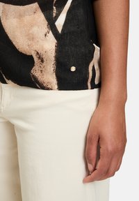 Czarna bluzka z abstrakcyjnymi beżowymi i brązowymi wzorami, teksturowana tkanina oraz mały złoty detal z logo, w połączeniu z jasnymi spodniami.