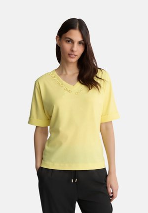 Top giallo a maniche corte con scollo a V e risvolti, caratterizzato da decorazioni lungo il colletto. Tessuto morbido, vestibilità rilassata, abbinato a pantaloni neri.