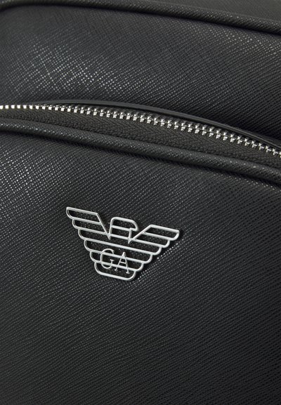 Svart texturerad ryggsäck med en silverlogo som har vingar och "GA." Dragkedjedetaljen avslöjar smidig funktionalitet.
