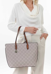 Šedá taška na rameno s diamantovým vzorem, hnědé kožené rukojeti a stříbrné kovové doplňky. Držena osobou v bílém tričku a neutrálních kalhotách.