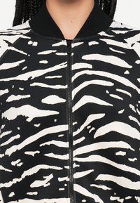 Veste bomber noire et blanche à rayures zébrées avec fermeture éclair, col côtelé et texture lisse. Présente des motifs audacieux et contrastés.