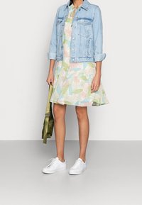 Vaaleansininen denimtakki kukallisella mekolla, jossa on vaaleanpunaisia, vihreitä ja sinisiä lehtikuvioita; yhdistetty valkoisiin lenkkareihin ja vihreään laukkuun.