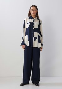 Button-up blouse in cream with navy abstract patterns. Wide-leg navy trousers. Smooth fabric, long sleeves with wide cuffs, and pointed black shoes.
