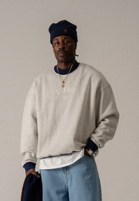 Helles graues Sweatshirt mit blauen Rippbündchen, mit aufgesticktem "BLKVIS NY". Getragen mit einem weißen, schichtartigen Hemd und blauen Jeans.