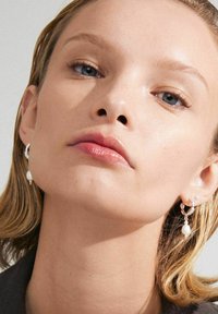 Silver hoops med en hängande pärla, tillsammans med en slät, naturlig läpp och glödande hud mot en neutral bakgrund.