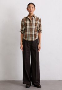 Chemise à carreaux marron et beige avec un col à boutons, manches retroussées ; assortie à un pantalon large marron foncé et des chaussures noires.