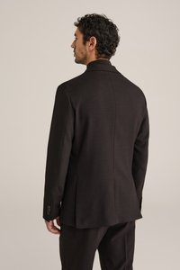 Homme aux cheveux bouclés portant un costume sombre sur mesure, debout et de profil sur un fond beige uni.