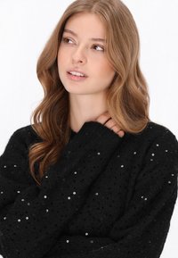 Fekete, textúrált kötött pulóver flitterekkel, laza fazonnal és hosszú ujjakkal. Az anyag otthonos megjelenésű, pöttyös mintázattal.