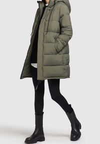 Piumino verde oliva con cappuccio, caratterizzato da cuciture orizzontali, tasche laterali e dettaglio con cerniera, indossato sopra leggings neri e stivali.