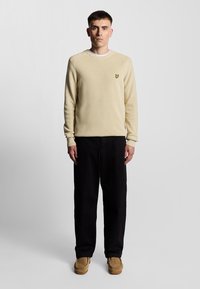 Beige strikket sweater med rund halsudskæring og ribstrikkede manchetter, der har et lille gult logo. Kombineret med sorte bukser med lige ben og brune sko.