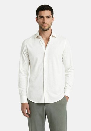 Uomo con camicia bianca a maniche lunghe abbottonata e pantaloni grigi in piedi davanti a uno sfondo chiaro e uniforme.