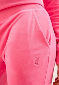 Rožinės vilnonės sportinės kelnės su minkštu paviršiumi, turinčios šoninį kišenę ir išsiuvinėtą logotipą kairiajame šlaunyje.