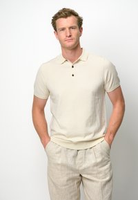 Ljusbeige polo skjorta med slim fit, korta ärmar och treknappsstängning. Bärs med ljusgrå linnbyxor.