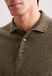 Primo piano di un uomo che indossa una polo in maglia verde oliva con due bottoni allacciati e un colletto ripiegato.