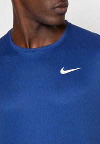 Man som bär en blå Nike t-shirt med en vit swoosh-logotyp på bröstet, tittar åt sidan mot en enkel bakgrund.