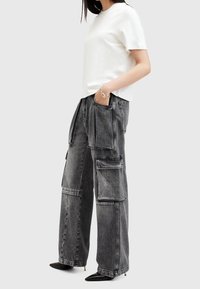 T-shirt blanc à manches courtes associé à un pantalon cargo en denim gris avec de grandes poches et une coupe décontractée. Des chaussures noires à bout pointu complètent la tenue.