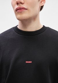 Μαύρη μπλούζα με ζιβάγκο από μαλακό ύφασμα, με μικρό κόκκινο λογότυπο "HUGO" στη θήκη. Ριμπ  λαιμόκ�οψη με απλό σχέδιο.