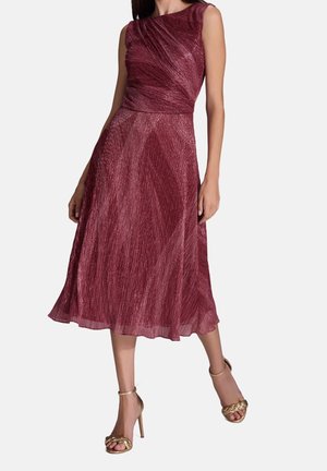 Vrouw die een mouwloze, glinsterende bordeaux midi-jurk met geplooide details en gouden sandalen met hoge hakken draagt, staand tegen een effen achtergrond.