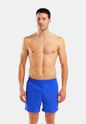 Uomo senza maglietta in piedi di fronte, indossa pantaloncini da bagno blu brillanti con vita elastica su uno sfondo bianco uniforme.