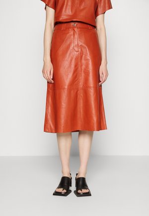 Kobieta ubrana w pomarańczową skórzaną spódnicę midi oraz top, do tego czarne sandały na obcasie z kwadratowym noskiem, stojąca na tle jednolitego tła.