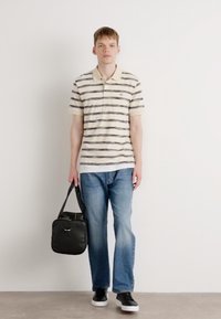 Tricou polo cu mânecă scurtă de culoare crem, cu dungi orizontale bleumarin și gri, purtat cu blugi albaștri deschiși și adidași negri; ținând o geantă neagră.