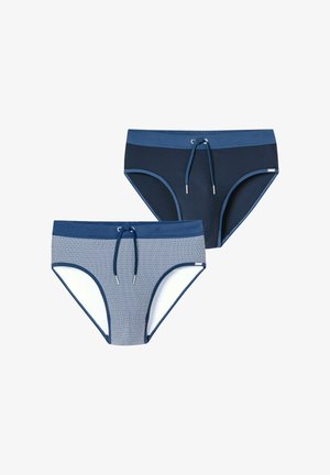 Jeune homme aux cheveux courts et bouclés, portant un maillot de bain bleu à motifs, debout devant un fond clair uni, ajustant ses mains.