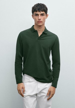 Young man with short dark hair wearing a dark green long-sleeve polo shirt and white pants, standing against a plain light background.