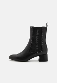 Botín negro hasta el tobillo con punta afilada, hecho de cuero suave. Presenta un panel elástico y texturizado con tachuelas y un pequeño tacón cuadrado.