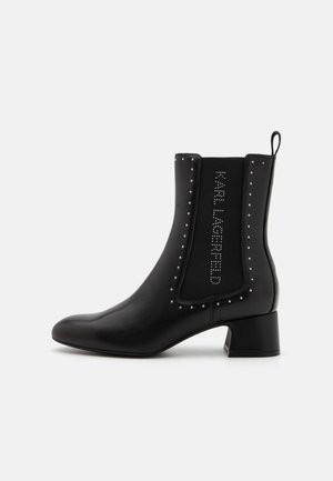 Stivale nero alla caviglia con punta affusolata, realizzato in pelle liscia. Presenta un pannello elastico e testurizzato con borchie e un piccolo tacco a blocco.